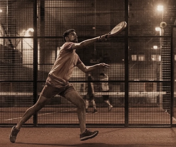 A medium shot shows a sweaty man lunging towards the center of a padel court, preparing to take a swing with an orange padel that is raised high. The background is a field with a wire-mesh fence that is dimly illuminated with several warm-toned light sources that create a bokeh effect.